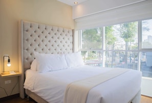 A neatly made bed with a tufted headboard, a bedside lamp, and a window with a view of trees and buildings.
