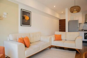 Una sala moderna con sofá y sillones, una obra de arte enmarcada, y una zona de cocina al fondo.