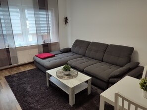 Moderni olohuone, jossa on harmaa kulmasohva, pieni valkoinen pöytä ja tummanharmaa matto.