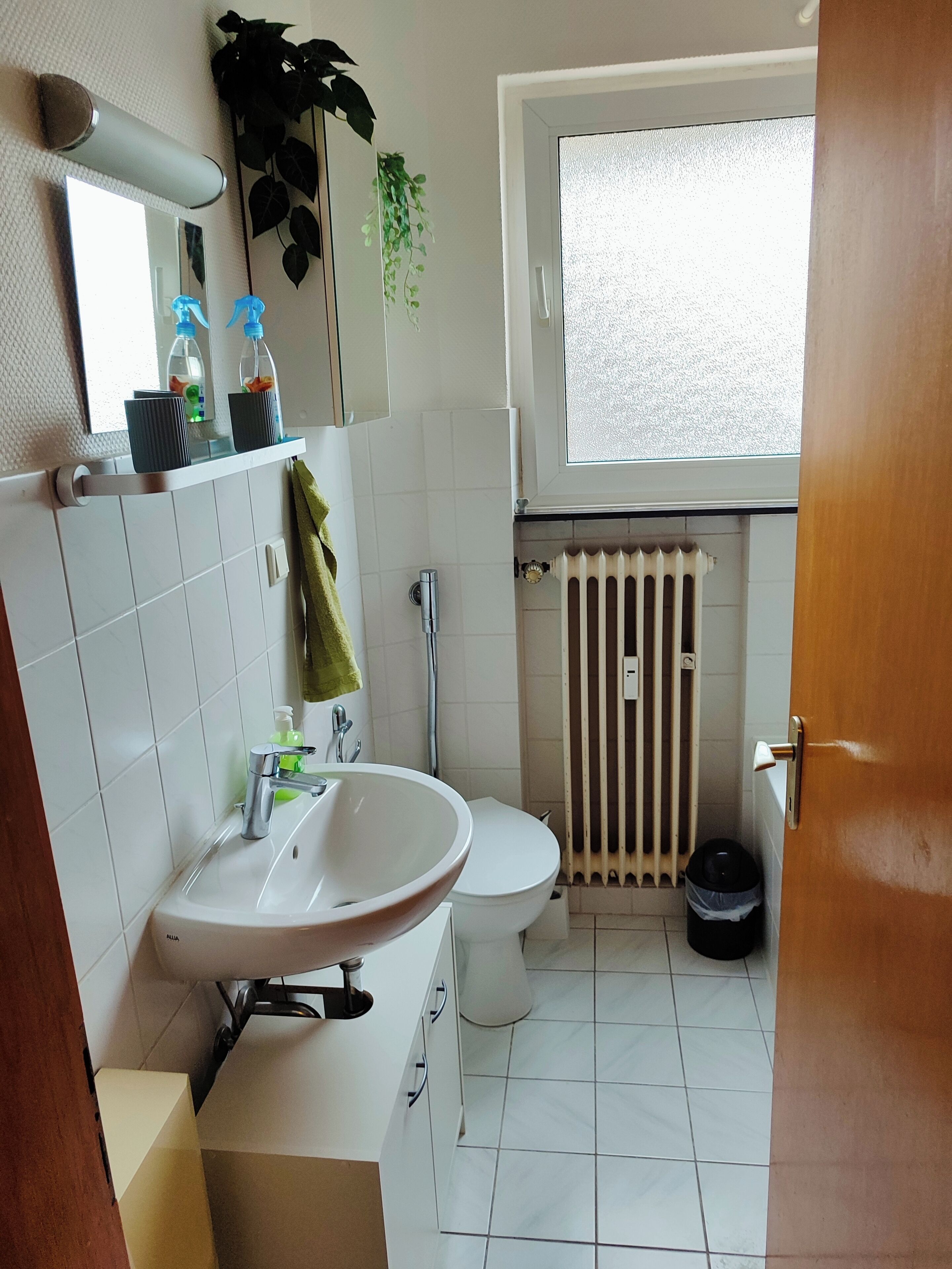 A bathroom with a white sink, toilet, and radiator. There is a window above the toilet and a towel hanging on the wall.