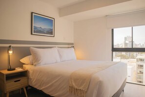A hotel room with a bed, a nightstand with a lamp, a framed picture on the wall, and a window with a city view. - w* | Enchanting 1BR w/ Balcony in Barranco (Barranco)