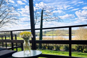 Un vase en verre contenant des fleurs, posé sur une table en verre sur un balcon d’où l’on a vue sur un lac et des arbres.