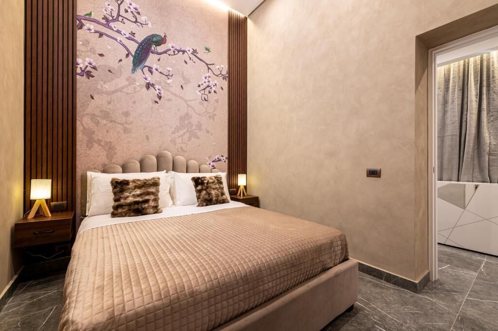 A modern hotel room with a large bed, a nightstand with a lamp, a wall mural of a bird on a branch, and a sliding door with curtains.