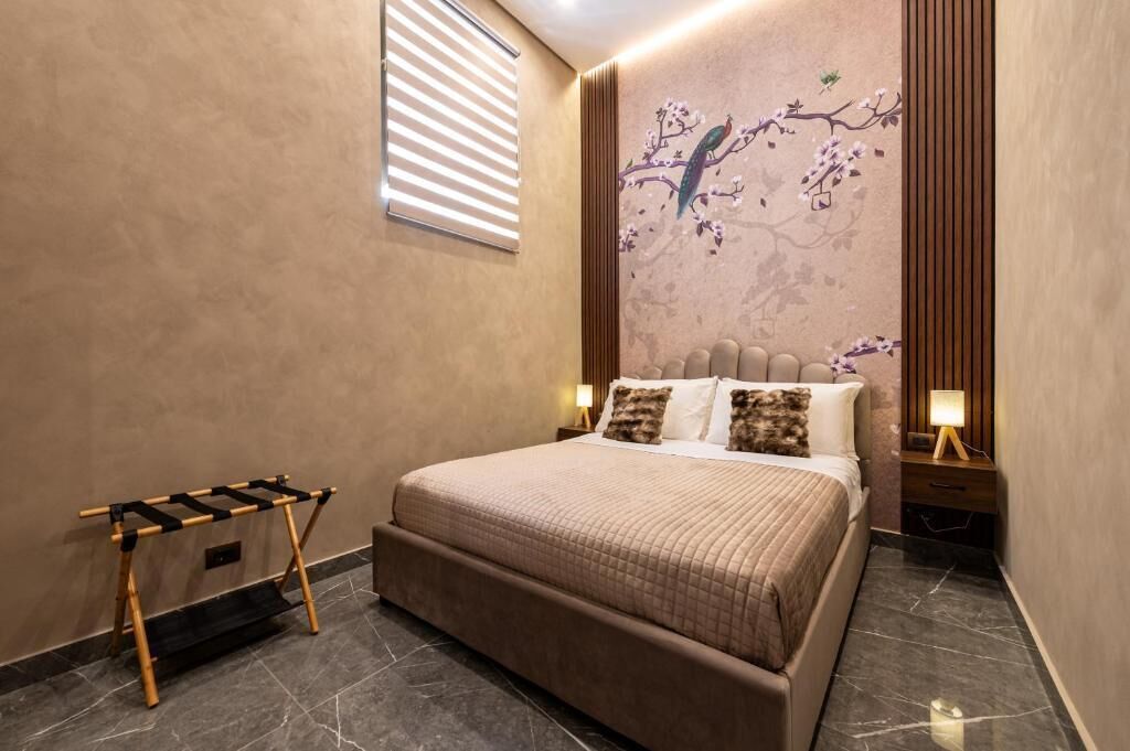 A modern bedroom with a large bed, a wooden side table, a wall-mounted shelf, and a window with blinds.
