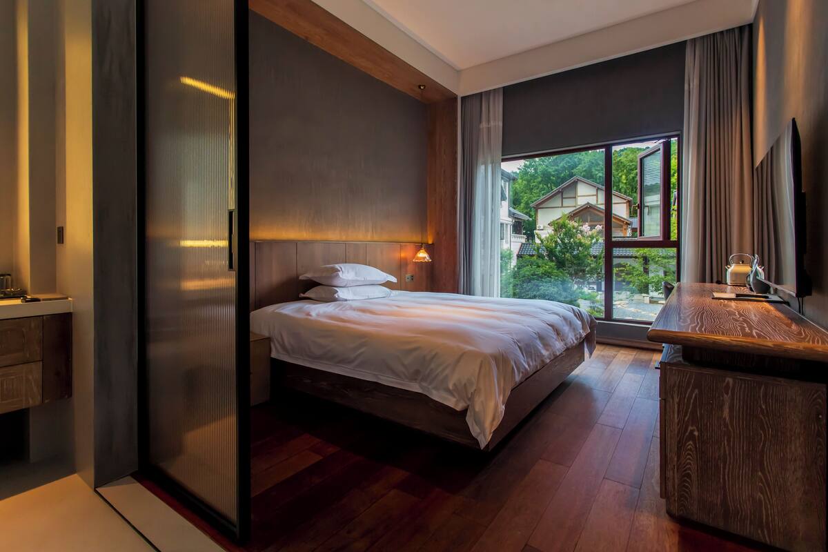 A modern bedroom with a large bed, a wooden desk, and a view of a garden through a large window.