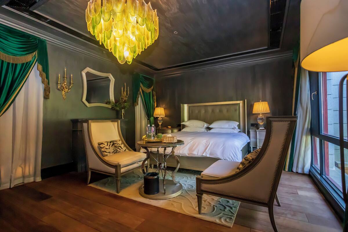 A hotel room with a bed, two chairs, a table, a chandelier, and a large window.