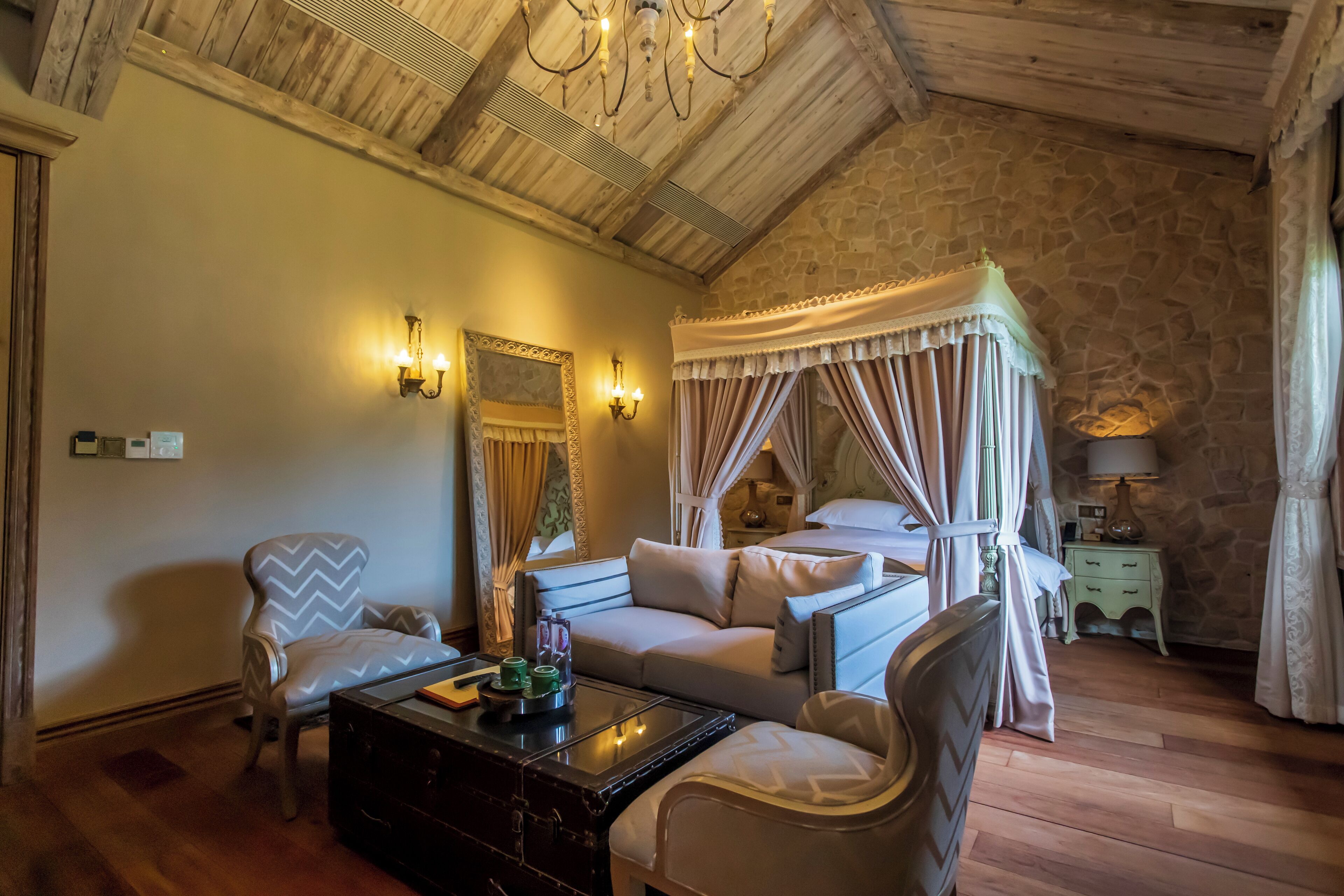 A bedroom with a four-poster bed, a sitting area with a sofa and armchairs, a nightstand with a lamp, and a stone wall.