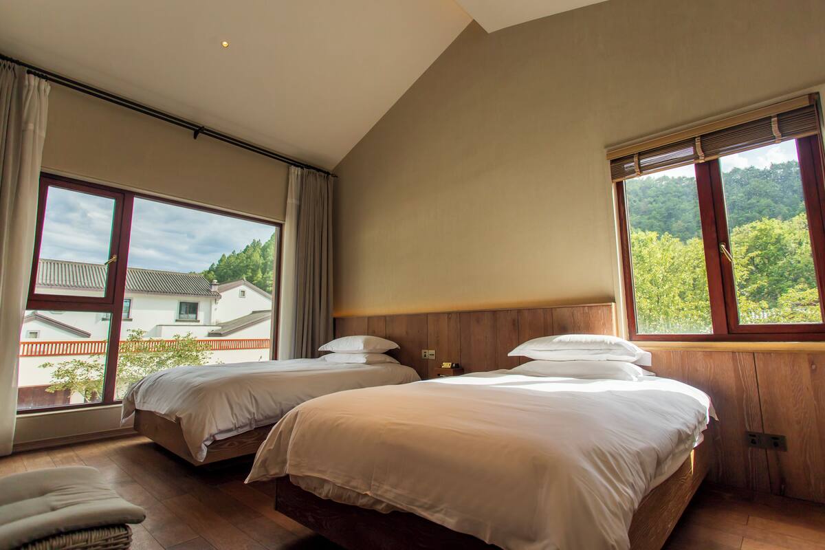 A bedroom with two beds, large windows, and a view of a house and trees.