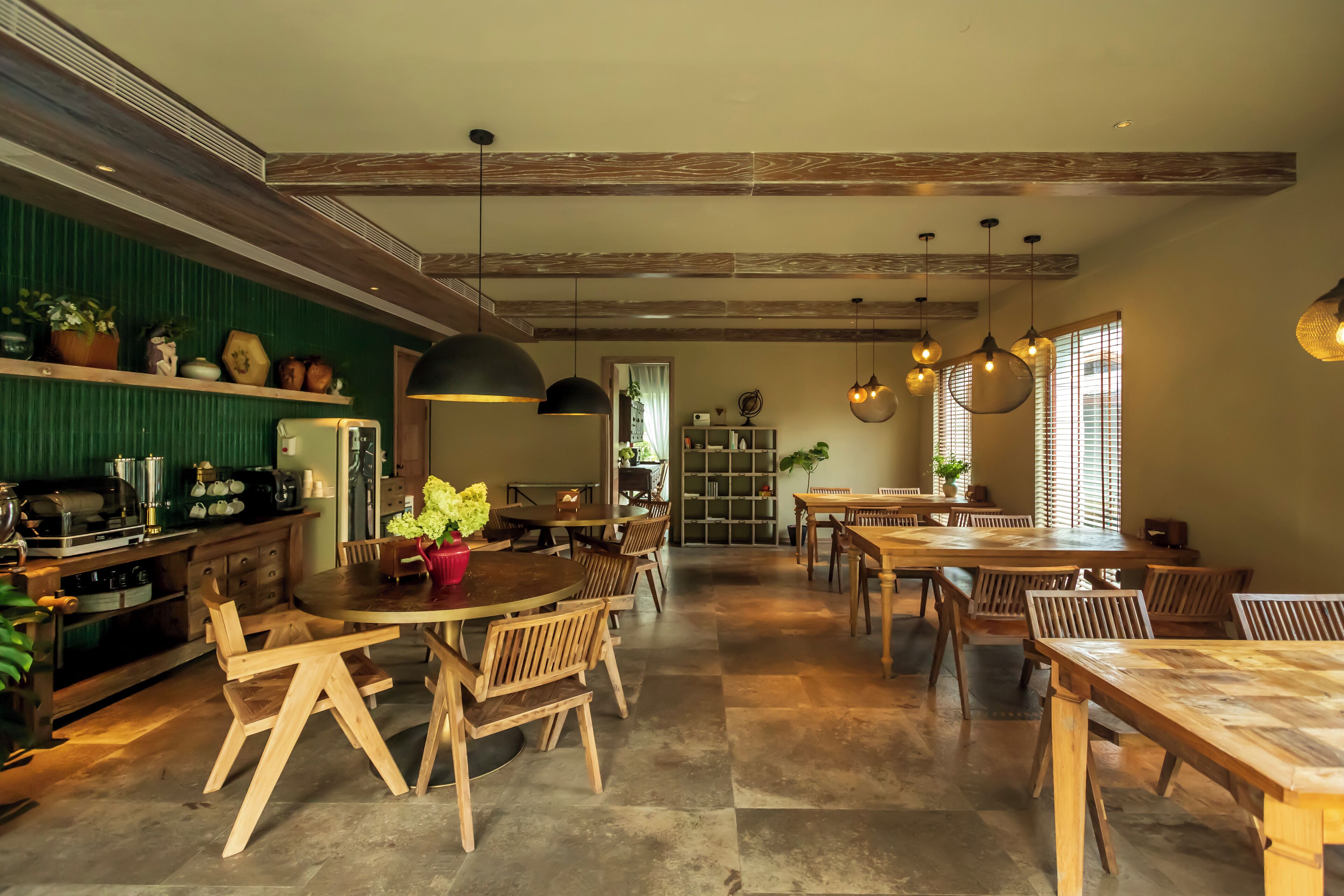 A spacious dining area with wooden tables and chairs, a kitchen area with shelves and appliances, and a view of the dining area through a doorway.