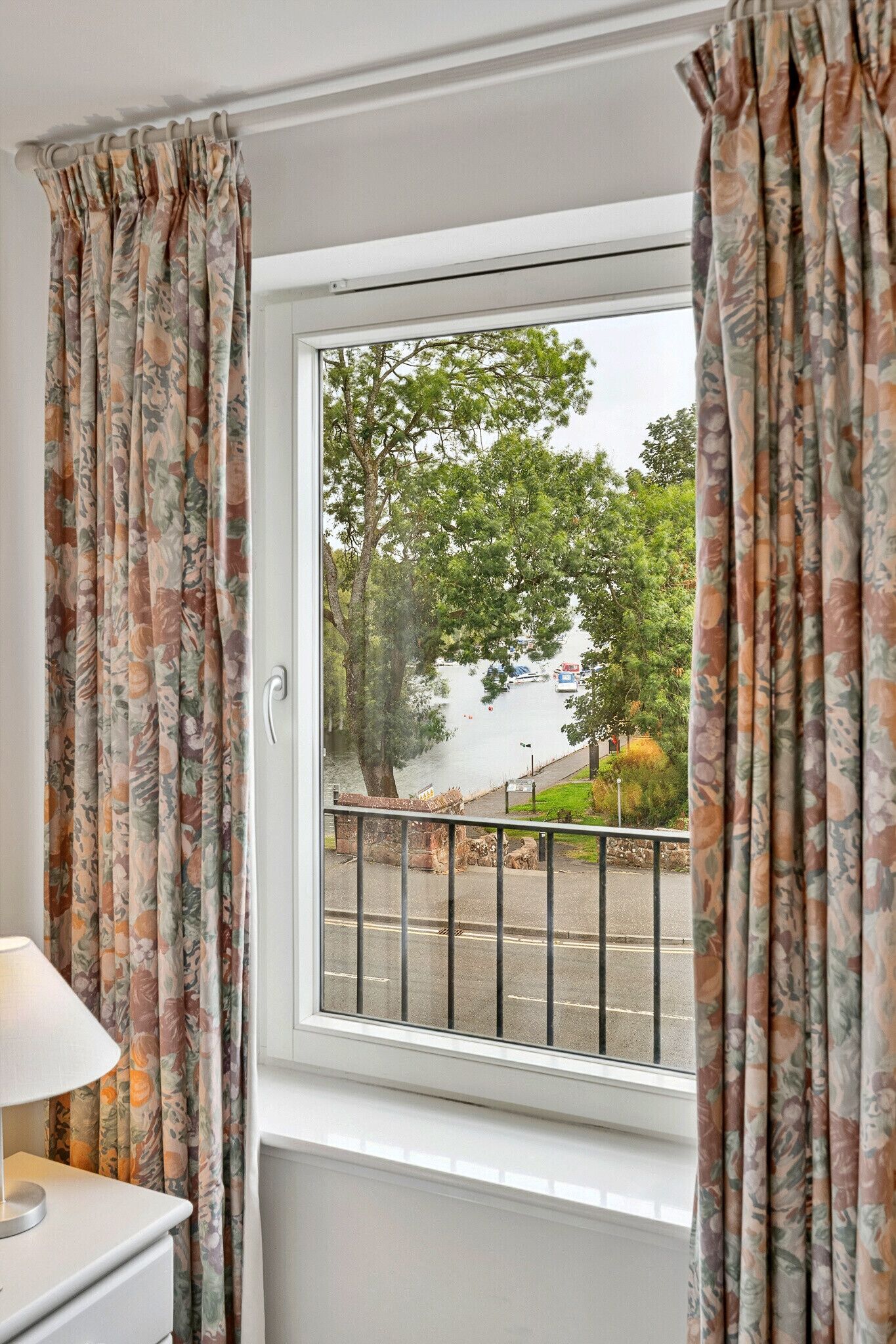 Habitación con ventana con cortinas florales, una mesita de noche blanca y vista a una calle arbolada y un cuerpo de agua.