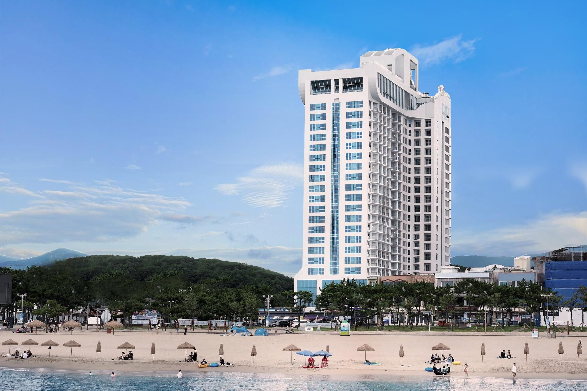 Un hotel alto dal design moderno, situato vicino a una spiaggia con ombrelloni e persone.