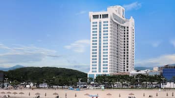 Un hotel alto dal design moderno, situato vicino a una spiaggia con ombrelloni e persone.