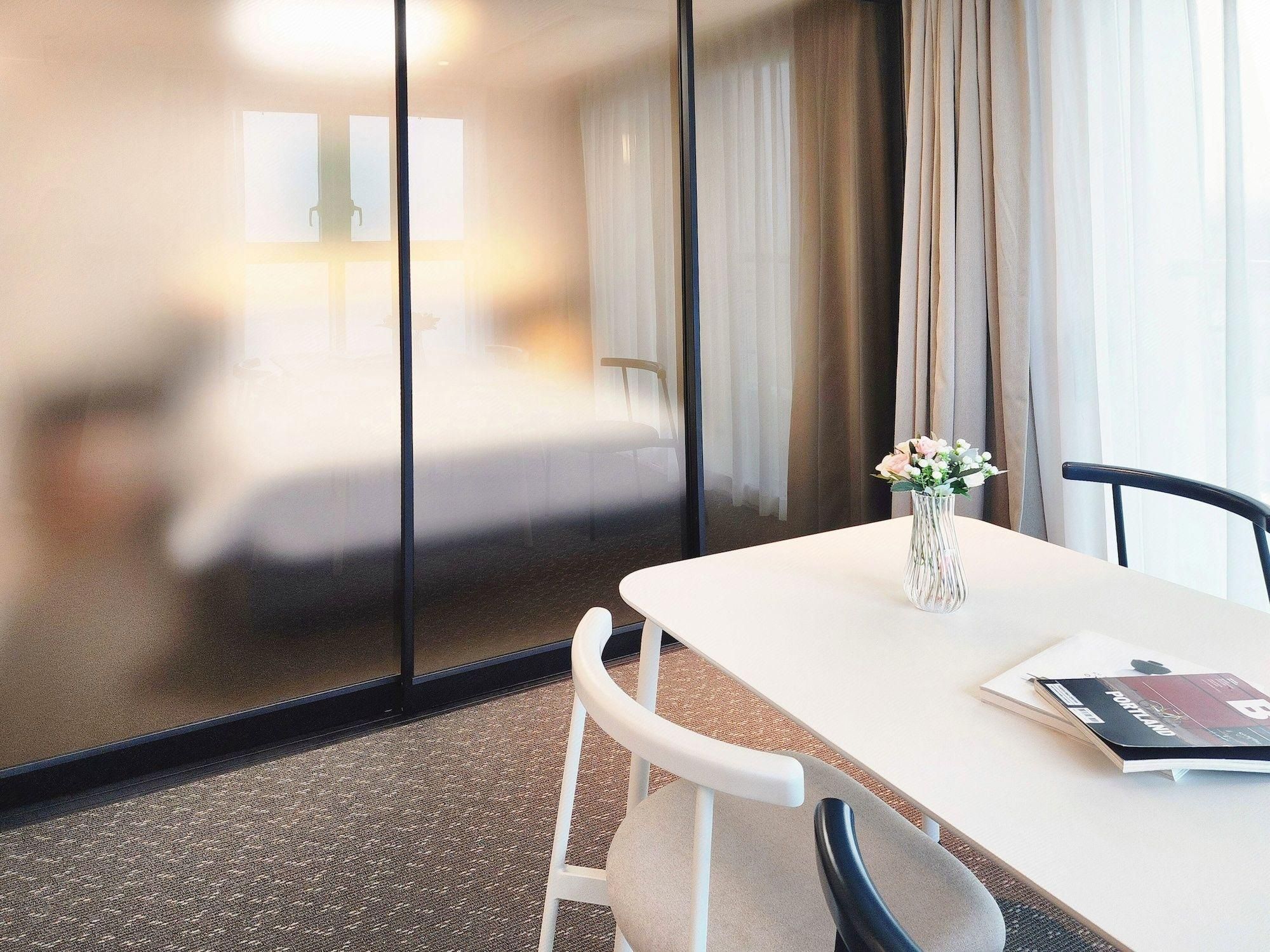 A hotel room with a white table, a vase with flowers, and a chair.