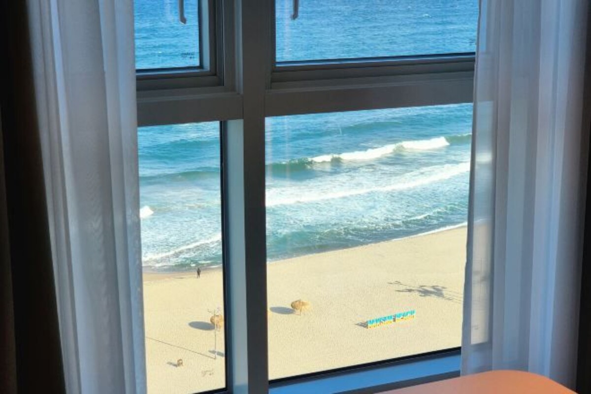 A window with a view of a beach, ocean waves, and a sandy shore.