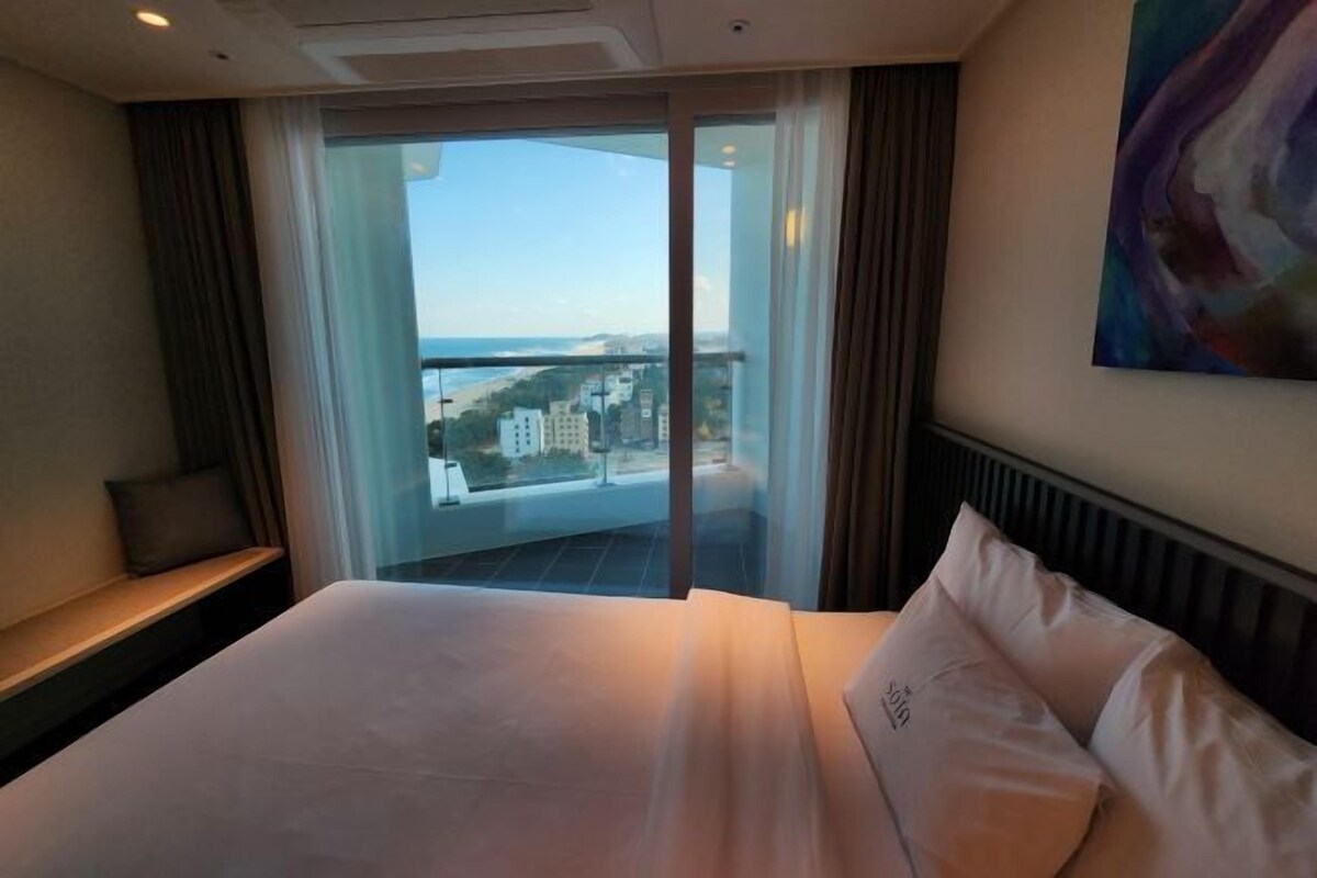 A hotel room with a bed, a window with a view of buildings, a painting on the wall, and a curtain.