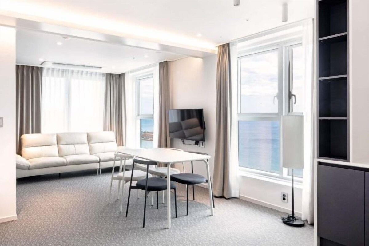 A modern hotel room with a sofa, a dining table with chairs, a flat-screen TV, and a large window.