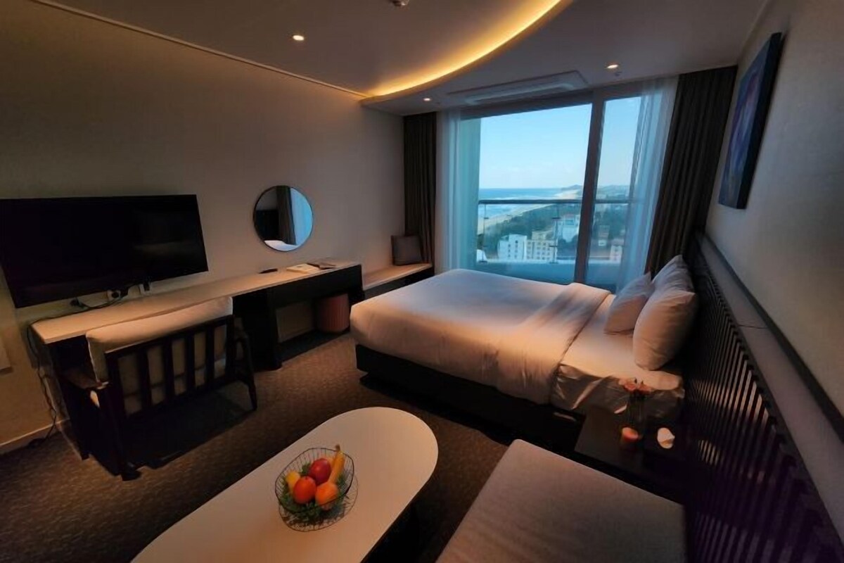 A modern hotel room with a large bed, a flat-screen TV, a desk, and a large window offering a city view.