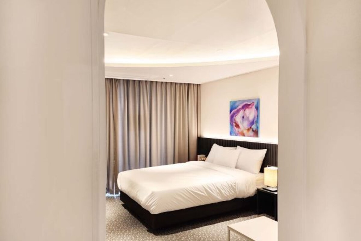A modern hotel room with a large bed, a nightstand, a painting on the wall, and a window with curtains.