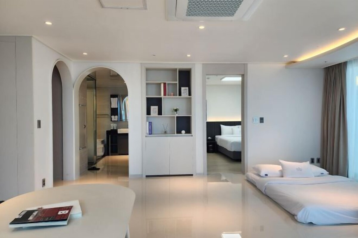 Een moderne hotelkamer met een bed, een bureau, een boekenkast en een boogvormige deuropening naar een andere kamer.