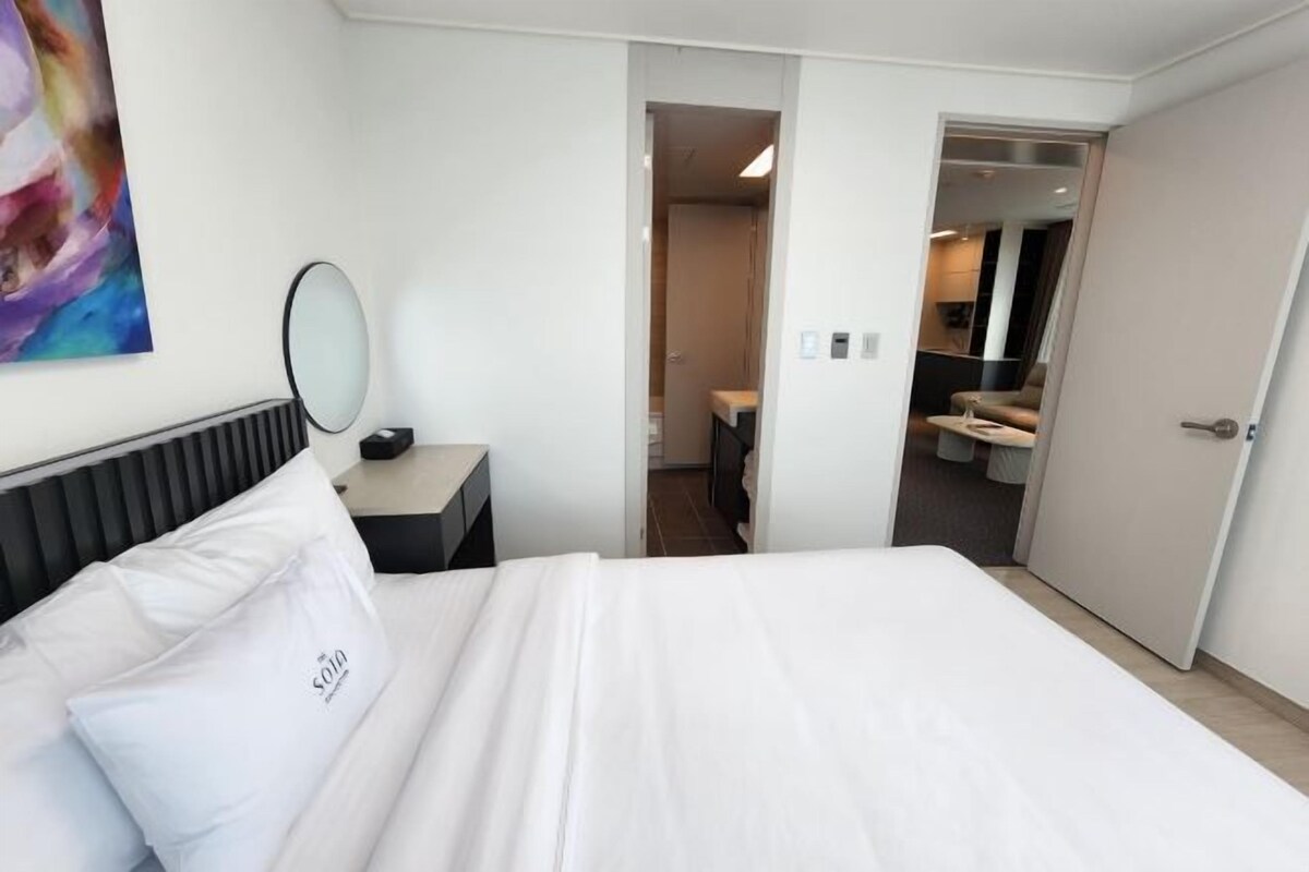 Een hotelkamer met een bed, een nachtkastje met spiegel, een schilderij aan de muur, en een open deur naar een badkamer.