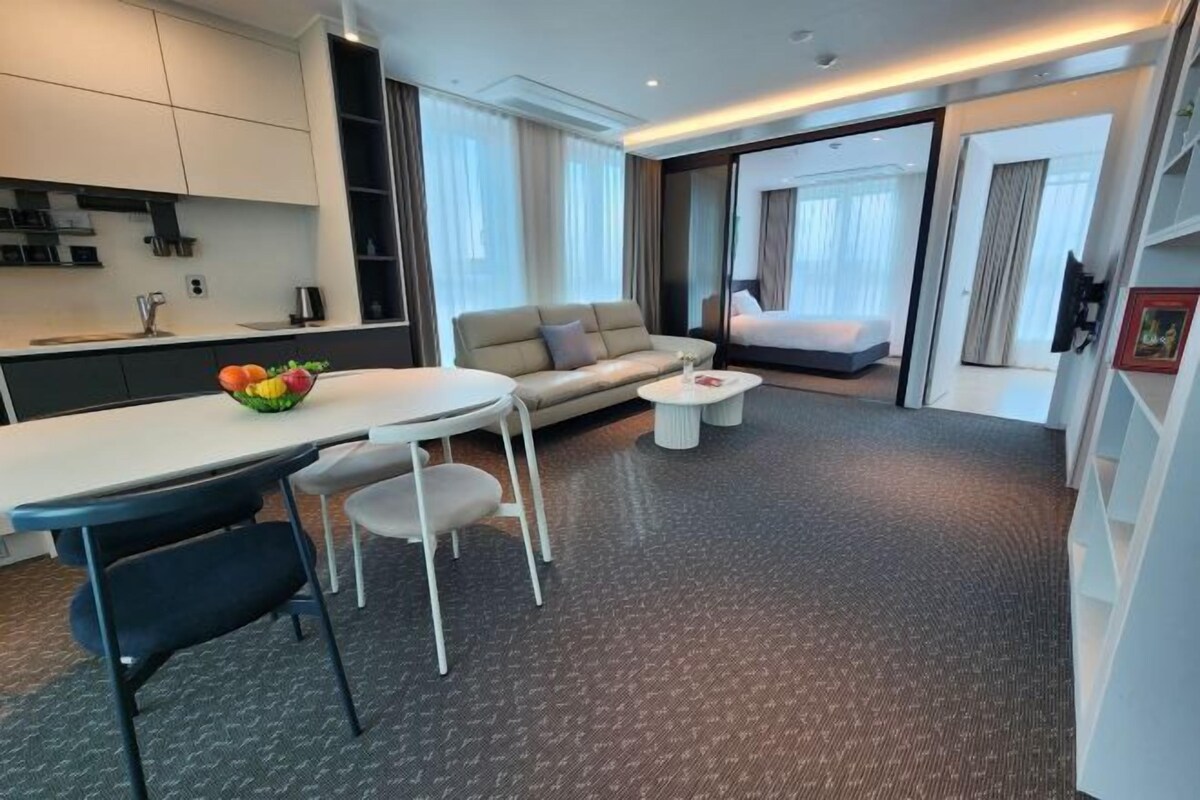 A modern hotel room with a sofa, a dining table with chairs, a small kitchen area, and a large bed.