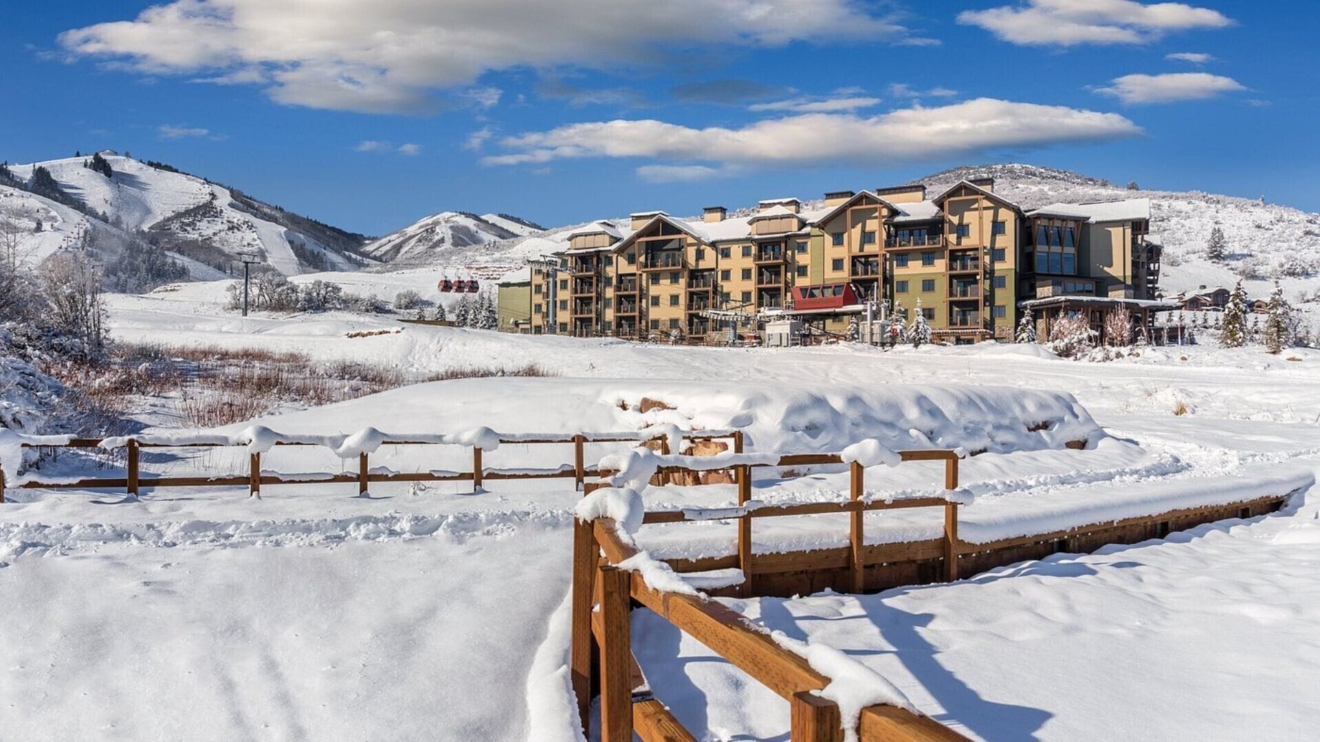 A snow-covered landscape with a wooden fence in the foreground and a multi-story hotel in the background.