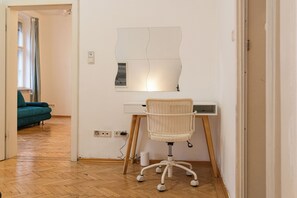 Ein Zimmer mit Holzboden, einem weißen Schreibtisch mit Korbstuhl, einem wandmontierten Spiegel und einem türkisfarbenen Sofa im Hintergrund.