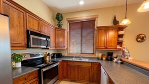 Una cocina con armarios de madera, microondas de acero inoxidable, horno, fregadero y ventana con persianas.