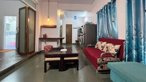A living room with a red sofa, a blue sofa, a dining table with chairs, a washing machine, and a kitchen area in the background. - Gaur Opulent Goa (Candolim)