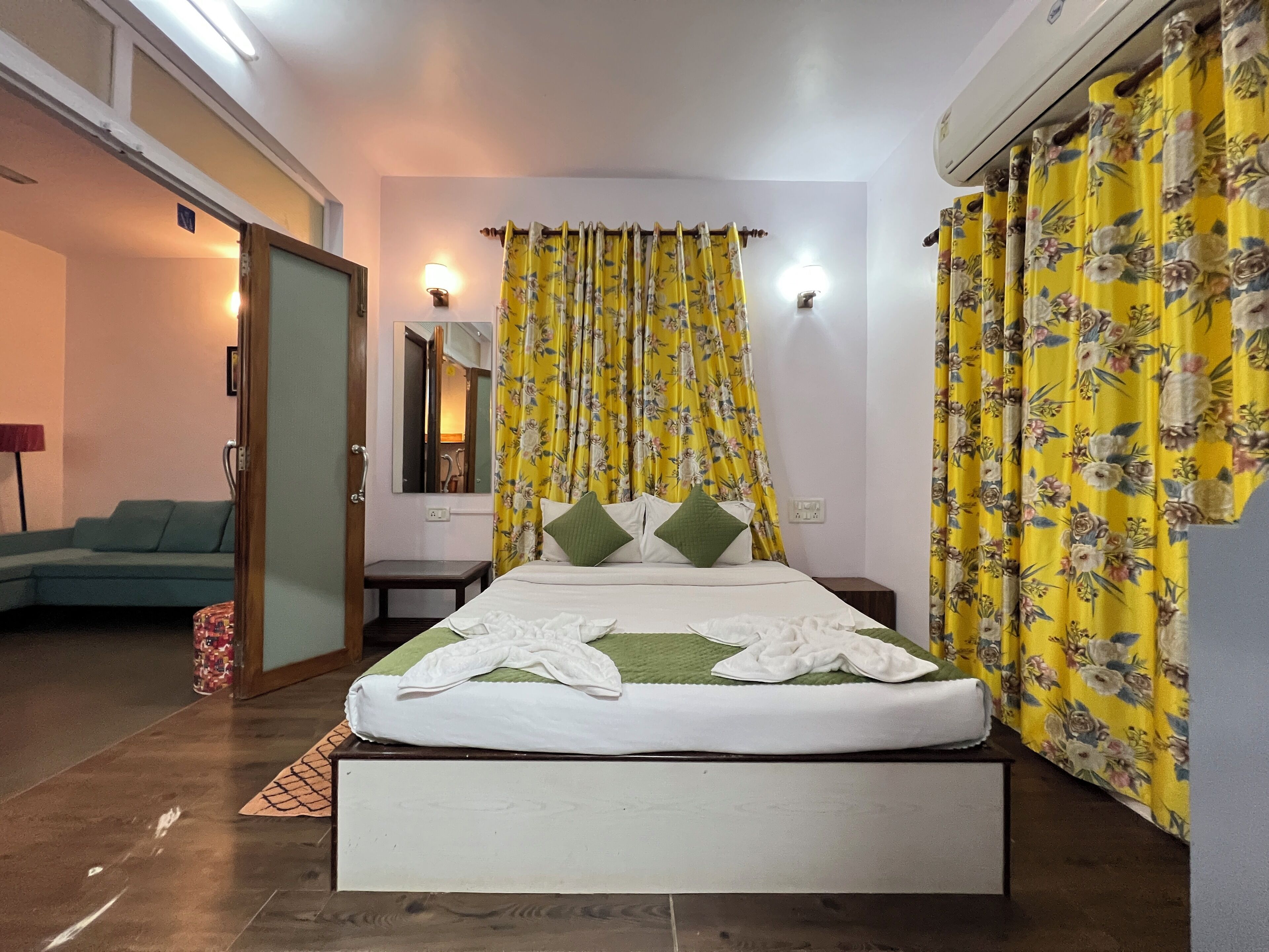 A bedroom with a bed, a bedside table, a nightstand, a mirror, and a window with yellow floral curtains.