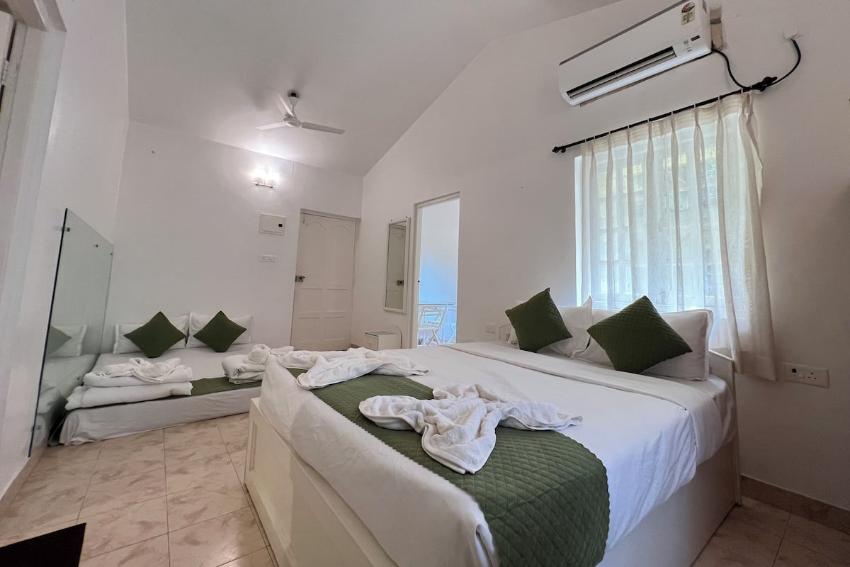 A hotel room with a large bed, a ceiling fan, an air conditioning unit, a window with sheer curtains, and a bathroom visible through an open door.
