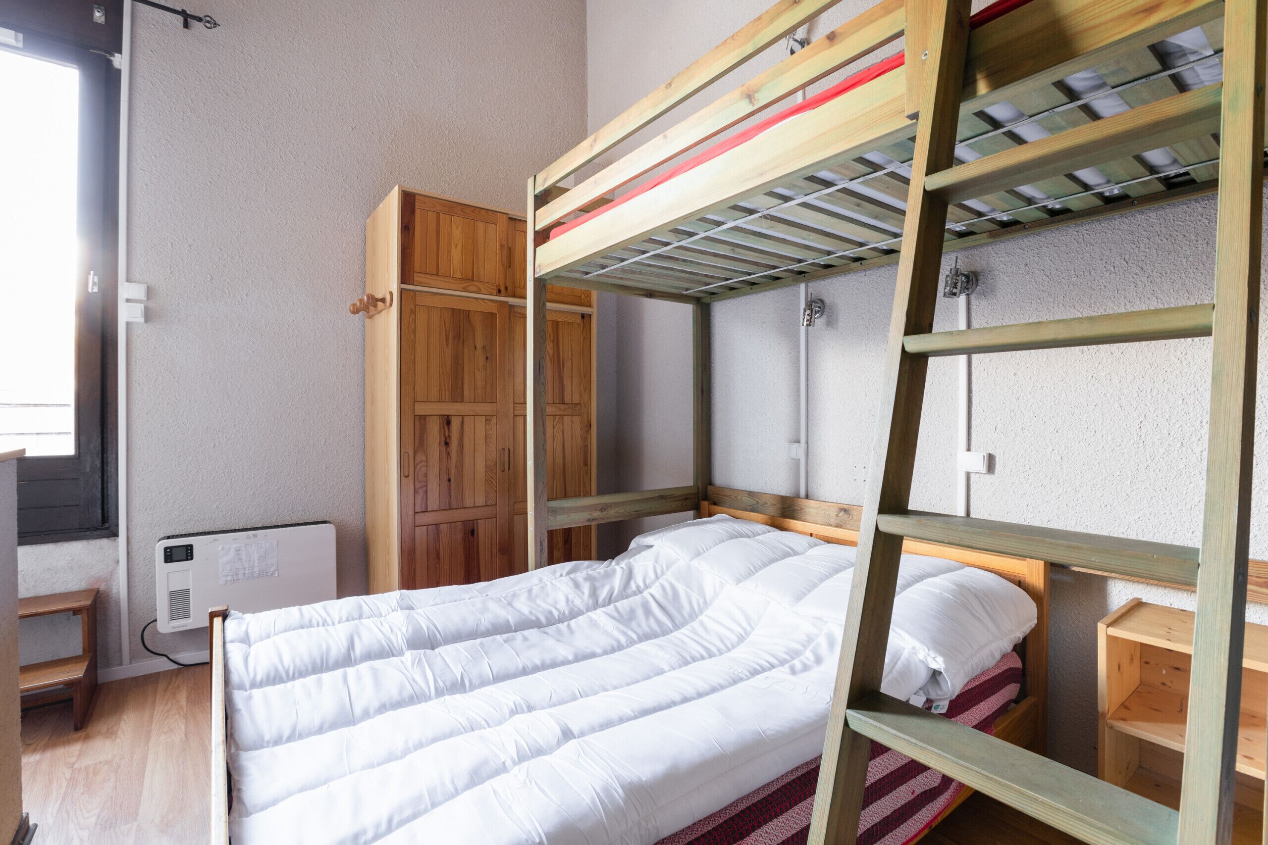 Un lit superposé avec une structure en bois, un matelas blanc et une couverture à rayures rouges et blanches. Contre le mur, on trouve une armoire en bois et un radiateur.