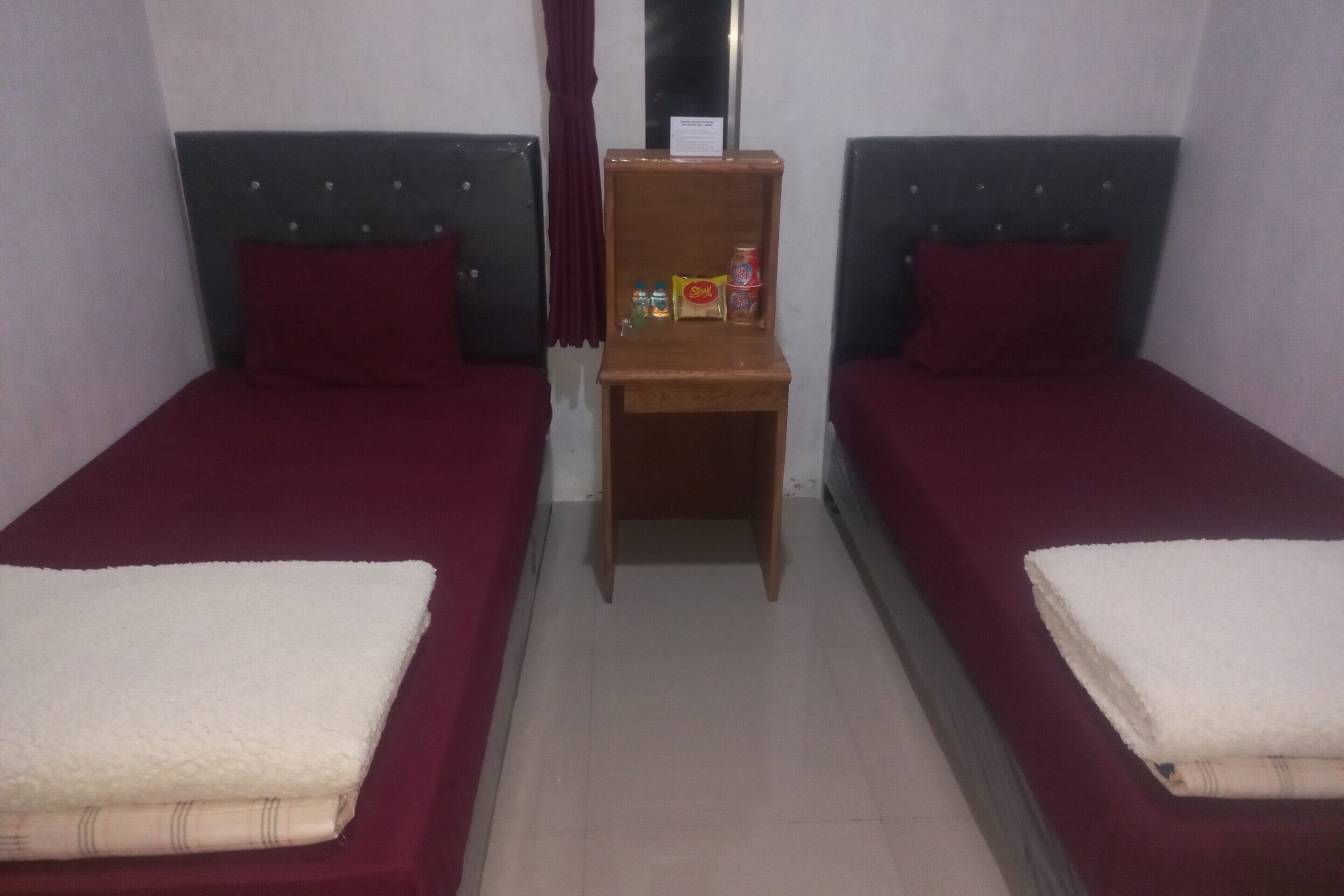 Ein Zimmer mit zwei Einzelbetten, jeweils mit roter Bettdecke und weißer Decke. Zwischen den Betten befindet sich ein Holzregal mit einer Flasche, einer Dose und einer Packung.