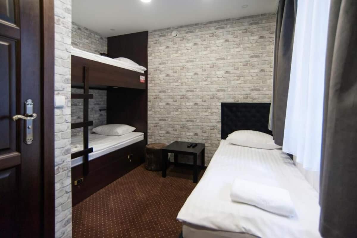A hotel room with a bunk bed, a single bed, a small table, and a brick-patterned wall.