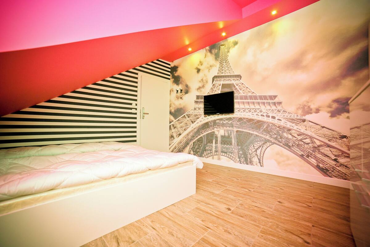 A bedroom with a bed, a striped headboard, a mural of the Eiffel Tower, and a TV mounted on the wall.
