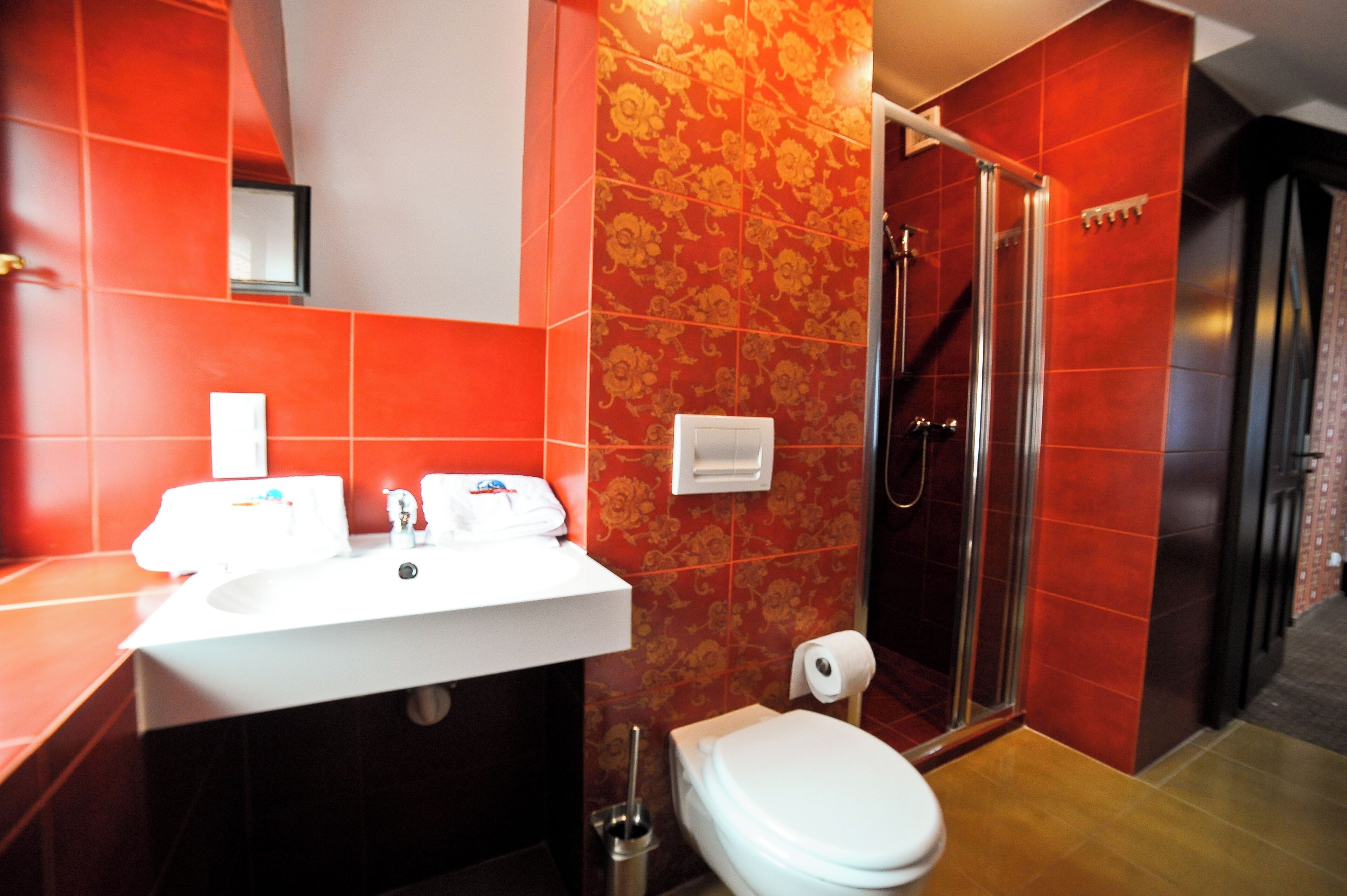 A bathroom with a white sink, toilet, and shower. The walls are tiled in red and orange, with a floral pattern.