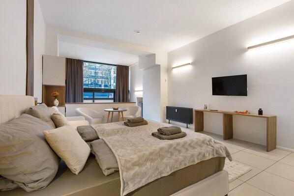 A modern hotel room with a large bed, a desk, a TV, and a view of the city.