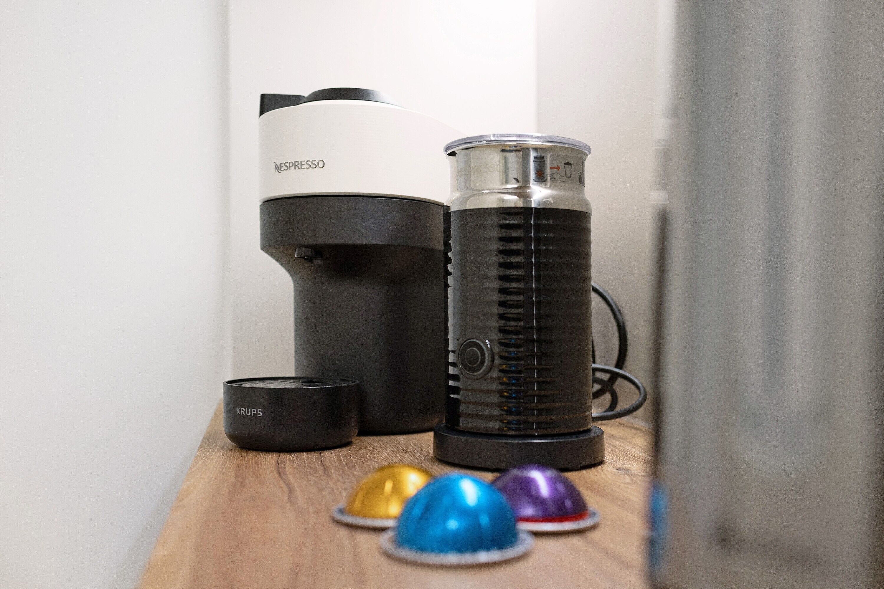 Una cafetera, un dispensador de agua y tres cápsulas de café sobre una superficie de madera.