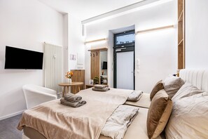 A hotel room with a bed, a chair, a TV, a radiator, a wooden door, and a small table with a vase of flowers. - City Loft Kurfürstendamm | Walk to Everything (3) (Berlin)