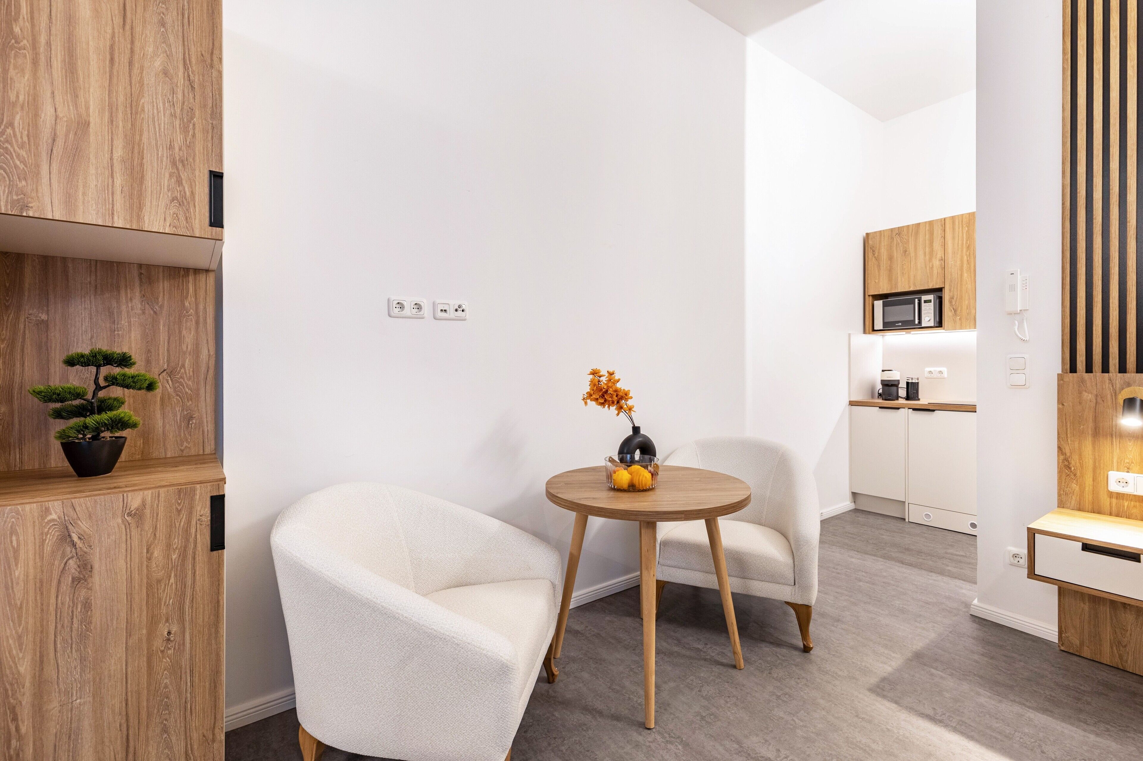 Ein modernes Zimmer mit einem runden Holztisch, zwei weißen Sesseln, einem Bonsai-Baum im Topf und einer kleinen Vase mit Blumen.