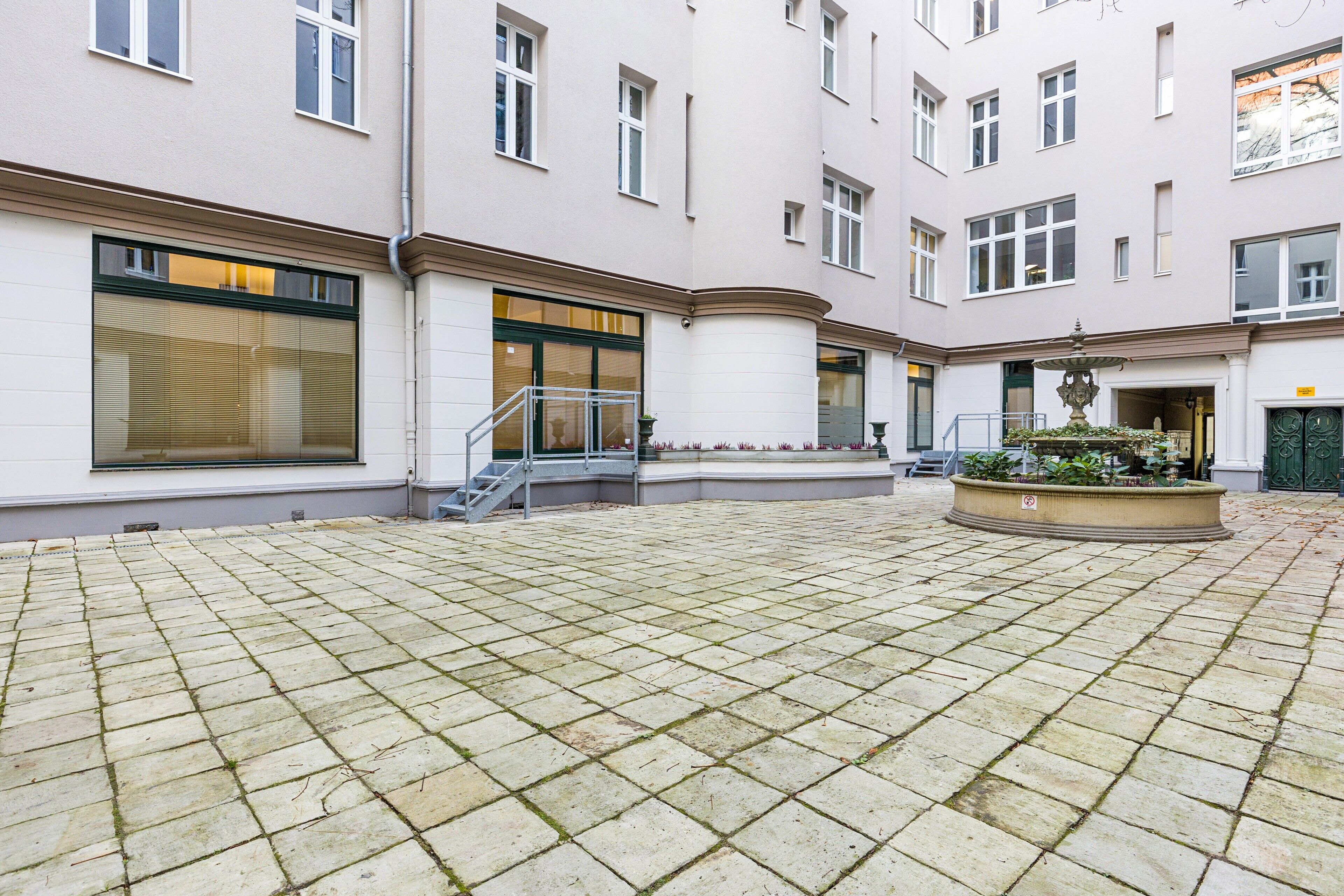 A courtyard with a tiled floor, a fountain, and a building with large windows and a door.