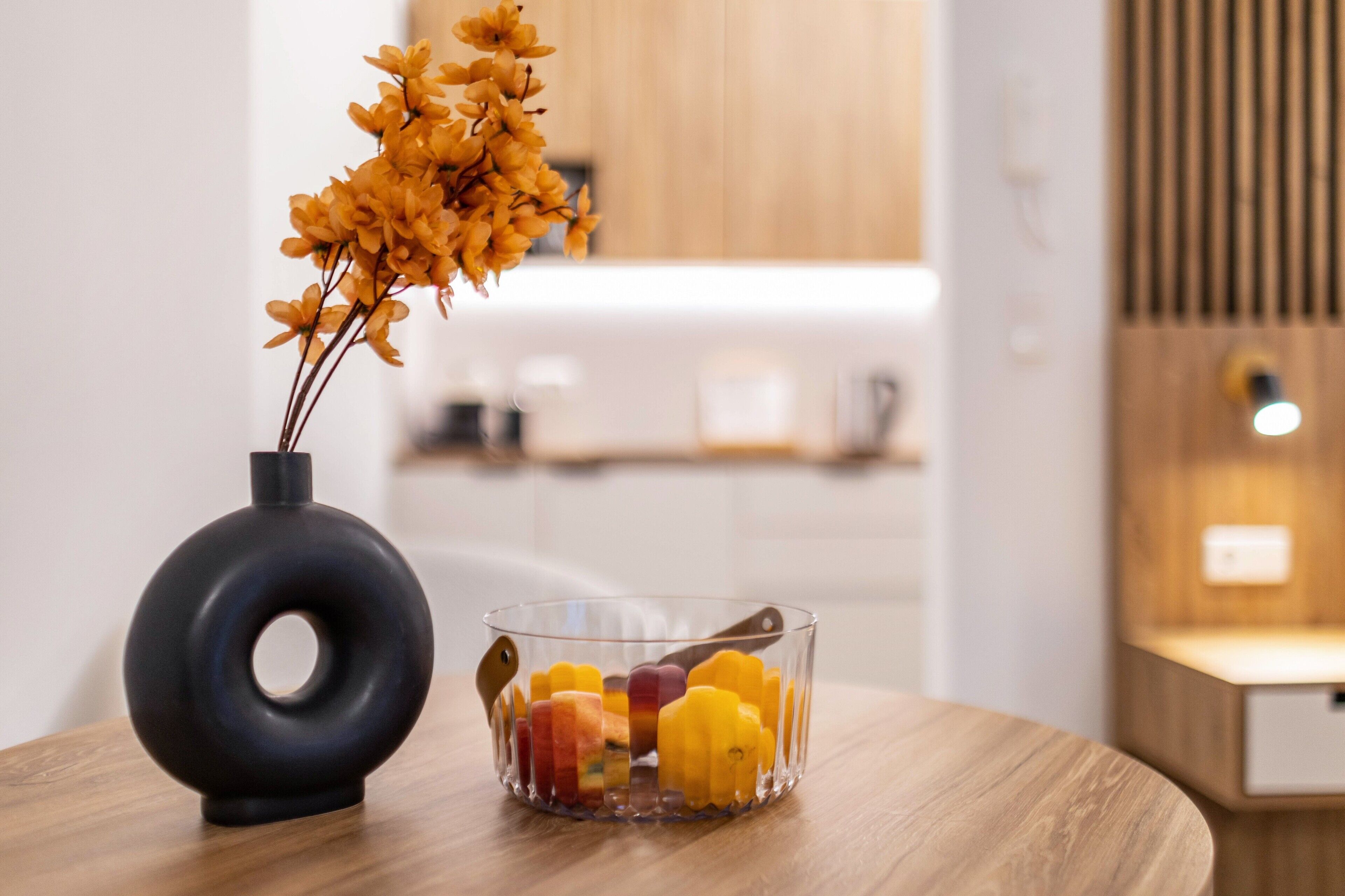 Eine moderne Küche mit einem Holztisch, einer schwarzen Vase mit Blumen und einer Glasbowl mit Obst.