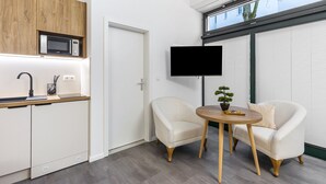 Moderni keittiö, jossa on mikroaaltouuni, pesuallas ja puinen kaappi. Seinälle on asennettu televisio, ja siellä on myös ovi sekä pieni pyöreä pöytä tuoleineen.