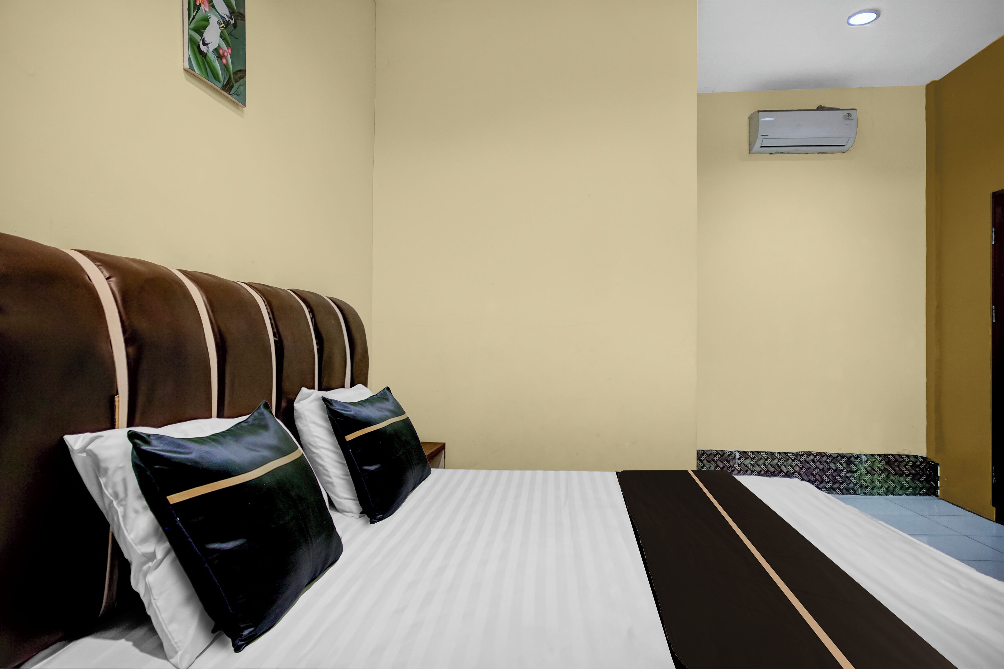 Habitación de hotel con cama, almohadas, cabecera, aire acondicionado de pared y un cuadro.