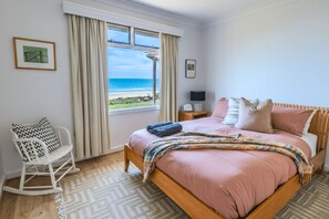 A bedroom with a bed, a rocking chair, a nightstand, and a window with a view of the beach.