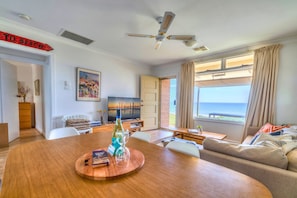 A spacious living area with a wooden dining table, a sofa, a TV, and a view of the sea through large windows. - Aldinga Stunning Sea View – 2BR Retro Vibe (Aldinga Beach)