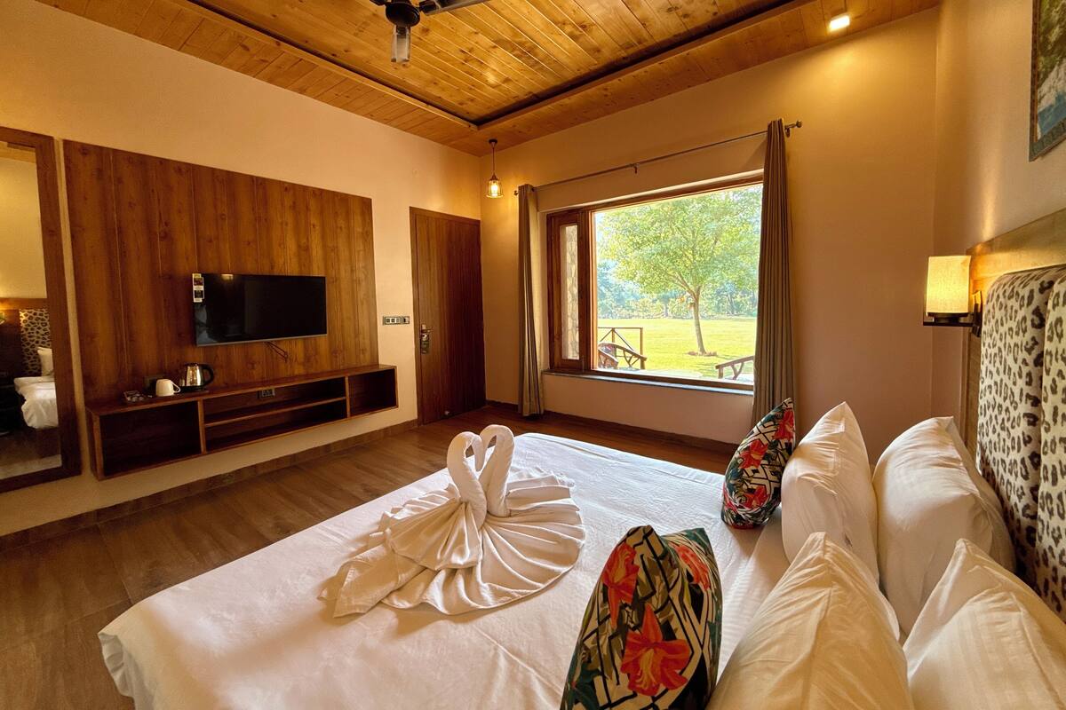 Une chambre d’hôtel avec un plafond en bois, un téléviseur à écran plat fixé au mur, un grand lit avec du linge de lit blanc, une fenêtre donnant sur un jardin et un ventilateur de plafond.