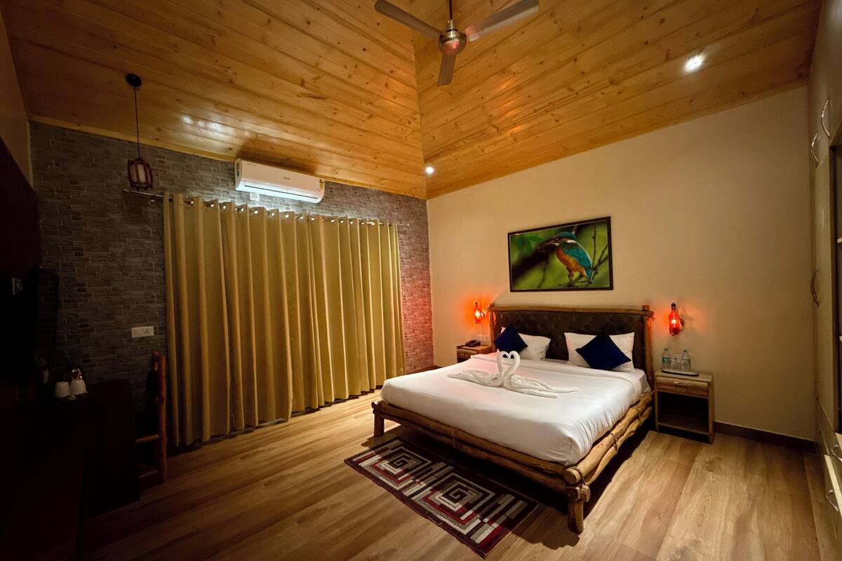 Une chambre avec un plafond en bois, un lit recouvert de draps blancs, une tête de lit ornée de coussins bleus, un climatiseur fixé au mur et un tableau représentant un oiseau.