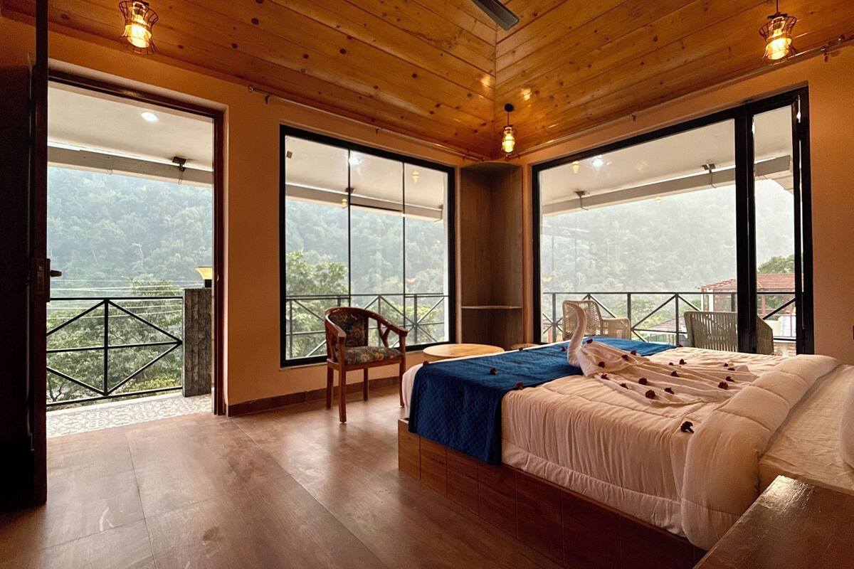 Une chambre avec un grand lit, une chaise et une petite table. De grandes fenêtres offrent une vue sur l’extérieur.