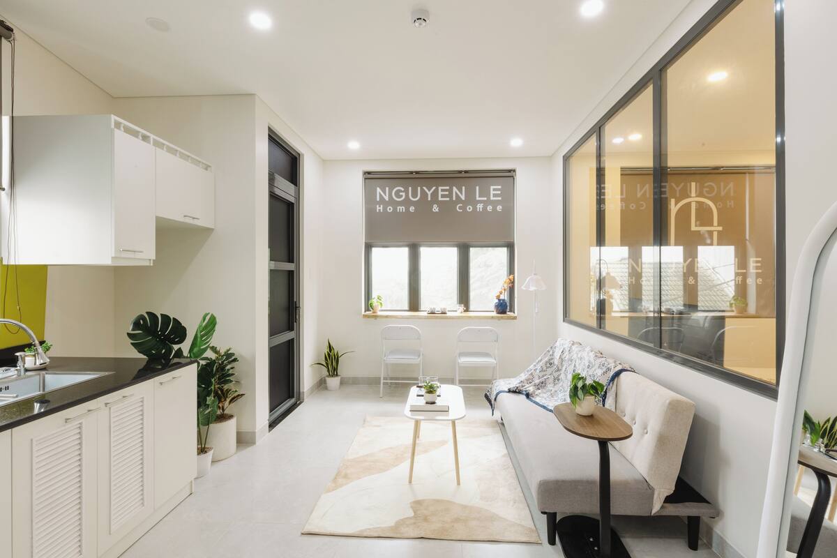 A modern kitchen with white cabinets, a sink, and a window with a view. There is a sofa and a small table with a plant.